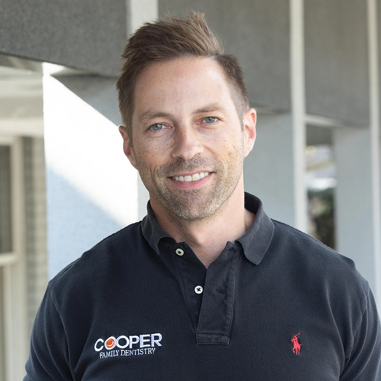 Male dental professional smiling at camera, wearing navy polo shirt with Cooper branding, indoor office setting.